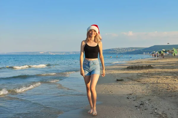 Девушка в шляпе Санта ходит по пляжу. Рождественские и новогодние праздники — стоковое фото