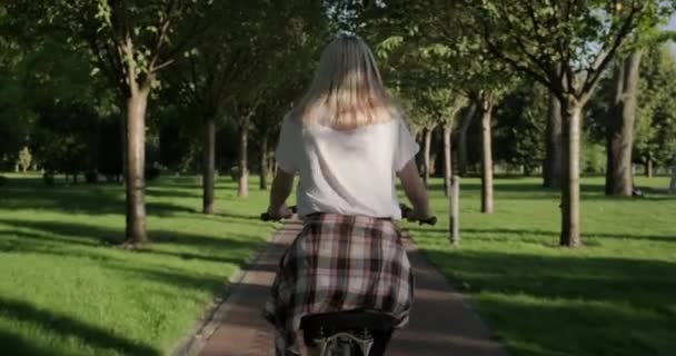 Hermosa chica rubia adolescente monta bicicleta en el parque, vista desde la parte posterior — Vídeos de Stock