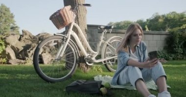 Güzel genç bir kadın akıllı telefondan video çekiyor. Parktaki çimlerde oturan bir kadın.