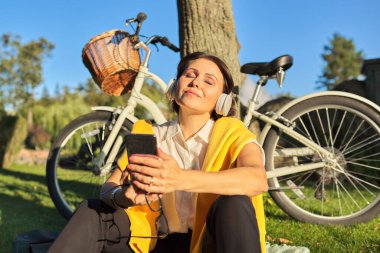 Parkta oturan, gözleri kapalı, kulaklık takan mutlu olgun bir kadın.