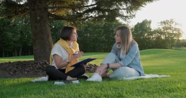 Female counsellor talking with young woman student teenager sitting on lawn in park on grass — Stock Video