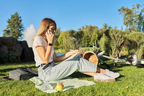 Młoda studentka siedzi w parku z podręcznikiem, dziewczyna studiuje za pomocą smartfona — Zdjęcie stockowe