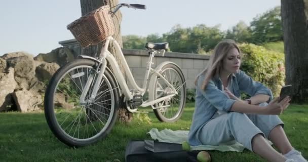 Красивая молодая женщина делает видеозвонок на смартфоне, женщина сидит на траве в парке — стоковое видео