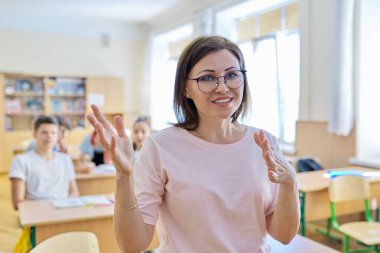 Sınıfta öğrencilerle konuşan orta yaşlı kadın öğretmen portresi.