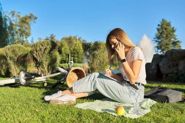Młoda studentka siedzi w parku z podręcznikiem, dziewczyna studiuje za pomocą smartfona — Zdjęcie stockowe