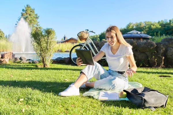 Teen girl student sitting with notebook digital tablet on green grass lawn in park