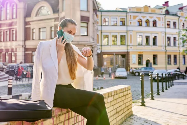 Mujer con máscara médica hablando en el teléfono celular al aire libre, mirando el reloj de pulsera —  Fotos de Stock