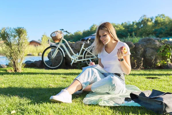 Menina bonita feliz estudante sentado na grama no parque com maçã, bicicleta — Fotografia de Stock