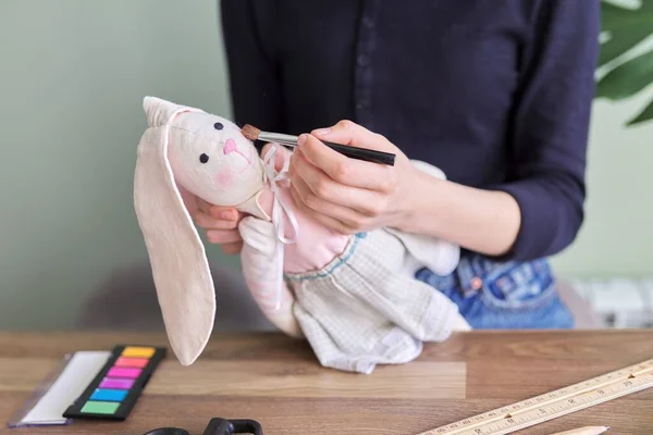 Muñeca de conejito hecha a mano en manos de una adolescente, la chica se pinta las mejillas con cosméticos — Foto de Stock