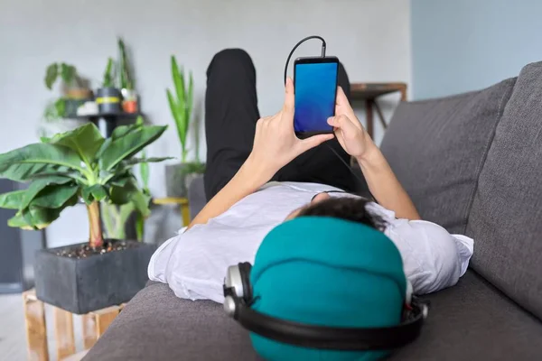 Smartphone schermo blu nelle mani di adolescente hipster sdraiato sul divano indossando cappello con cuffie — Foto Stock