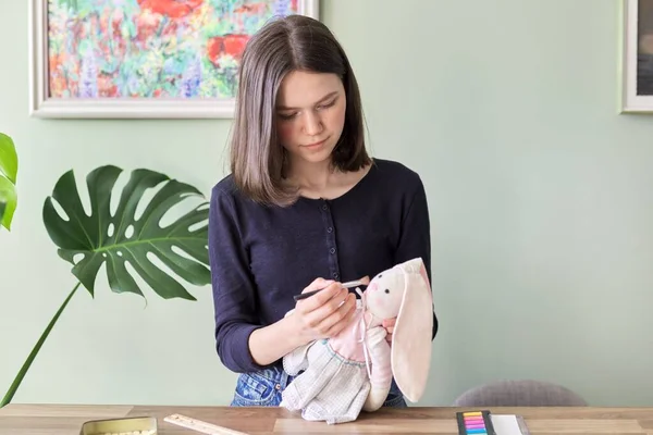 Tiener meisje naaien bunny pop speelgoed, zitten thuis aan tafel, tekent roze wangen met oogschaduw — Stockfoto