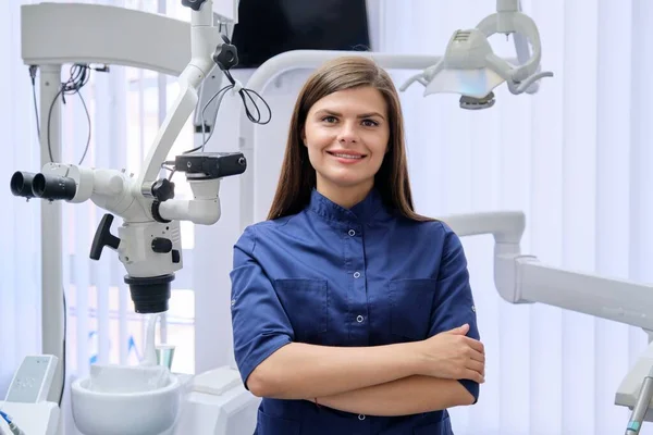 Portret van jonge zelfverzekerde vrouwelijke tandarts met gevouwen handen in tandartspraktijk. — Stockfoto