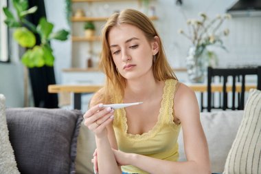 Evde oturan termometreye bakan üzgün genç kız. Yüksek sıcaklıkta mevsimsel viral hastalıklar, hastalık, sağlık gençliği, ergenlik kavramı