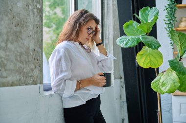 Olgun, üzgün, bunalımlı, stresli bir kadın, pencerenin yanında duran zihinsel ve fiziksel acı. Sorunlar yalnızlık, baş ağrısı hormon stresi, orta yaşlı kadınların sağlığı