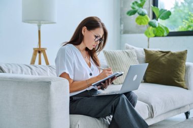 Orta yaşlı ciddi bir kadın evdeki bilgisayarlı not defteriyle kanepede çalışıyor. Kendine güvenen olgun bir kadın not alıyor, iş ödemeleri üzerine serbest meslek eğitimi alıyor.