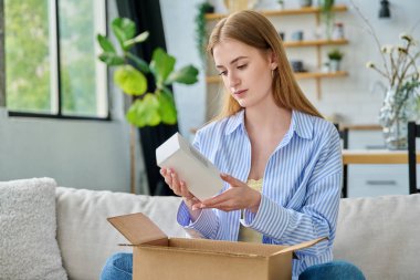 Genç, mutlu, karton kutuları internet siparişiyle açan, cilt bakımı parfümlü ev yapımı kimyasallar alan, evinde kanepede oturan bir kadın. E-alışveriş, e-market, teslimat servisi.