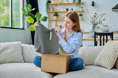 Karton kutuda paketleri açan mutlu genç bir kadın sipariş edilmiş kıyafetlere bakıyor, kanepede oturuyor. E-alışveriş, e-market, teslimat servisi, online kıyafet siparişi.