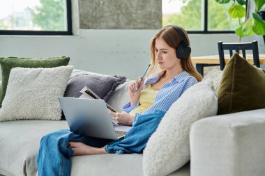 Kulaklıklı, dizüstü bilgisayarlı genç kız ders kitabı ile evdeki kanepede oturmuş online video dersi seyrediyor. Eğitim eğitimi teknolojileri gençlik iletişiminde eğlence amaçlı çalışır