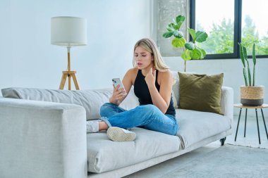 Mutsuz, üzgün, genç bir kadın akıllı telefon ekranına bakıyor. Oturma odasındaki kanepede oturuyor. Olumsuz duygular, stres, depresyon, akıl sağlığı, sorunlar kötü haberler