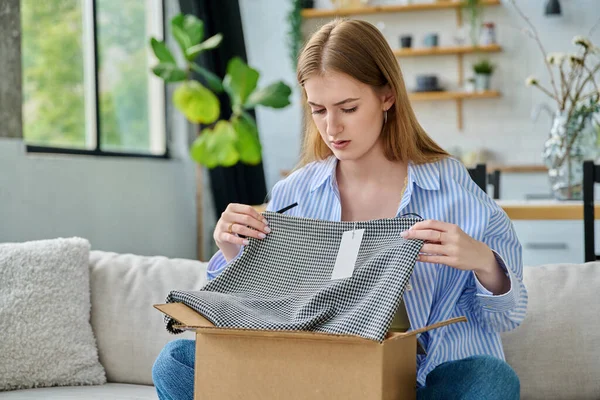 Karton kutuda paketleri açan üzgün genç bir kadın sipariş edilmiş kıyafetlere bakıyor, kanepede oturuyor. E-alışveriş, e-market, teslimat servisi, online kıyafet siparişi.