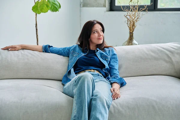 Oturma odasındaki kanepede oturan güzel, mutlu esmer kadının portresi. Gülümseyen, gevşemiş, çekici 30 'lu yaşlarda bir kadın. Güzellik yaşam tarzı rahatla rahat rahat mutluluk, kadın konsepti