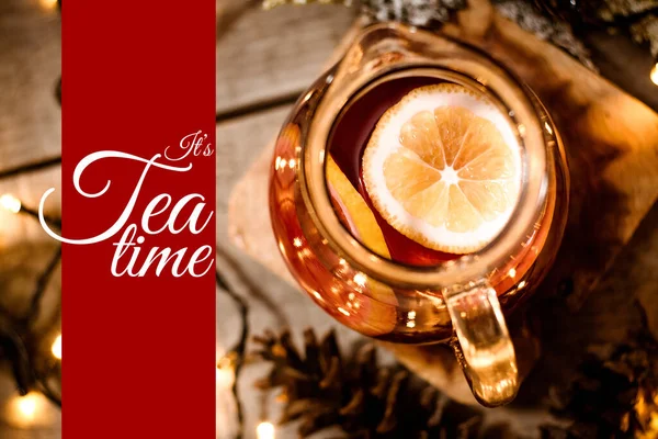 Perspective above of a carafe with fruit tea and a piece of lemon in it on a wooden table with bokeh and christmas lights with text It\'s Tea time