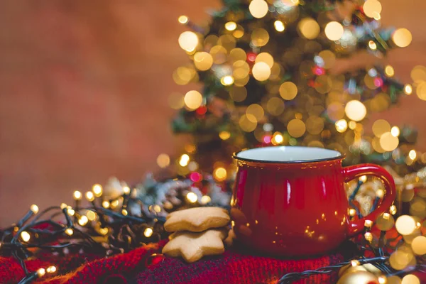 Kırmızı fincan sıcak kahve, Noel ışıkları, taze kurabiyeler ve Noel ağacı.