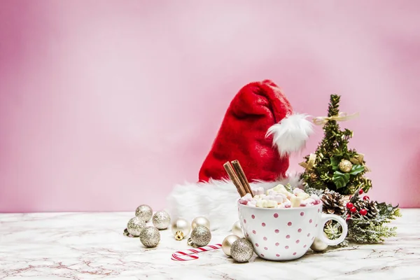Marşmelovlu pembe bir kupa, Noel ağacı, süsler ve mermer ve pembe arka planda Noel Baba şapkası.