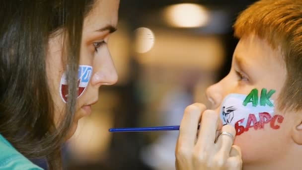 Замедлительная женщина рисует аква-макияж на щеке маленького мальчика — стоковое видео