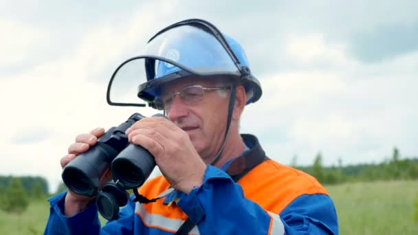 Engineer in helmet watches in binoculars high voltage line in field — Stock Video