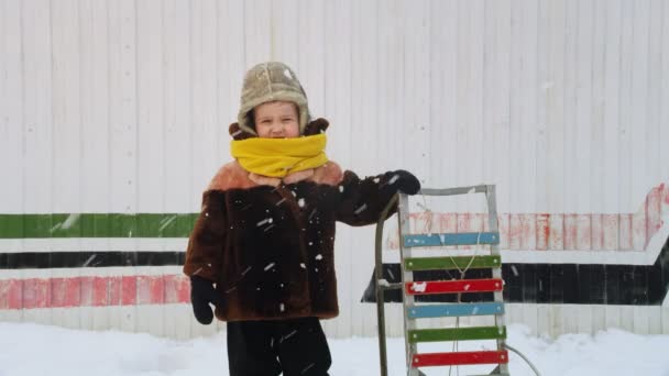 Милий хлопчик в зимовому одязі з саней стоїть на снігопазі — стокове відео