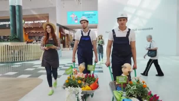 Mulher com brochuras e homens com plantas em carrinhos no mercado — Vídeo de Stock