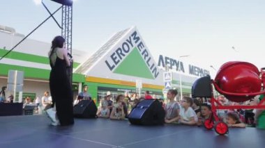 Siyah elbiseli şarkıcı süpermarketin yanındaki çocuklara gösteri yapıyor.