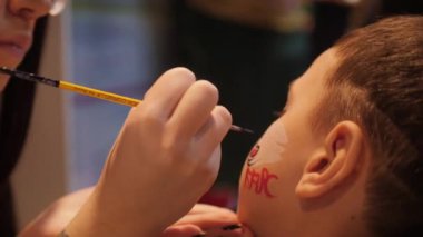 Kadın, küçük çocuğun yüzüne fırçayla hokey takımı sembolü çiziyor.