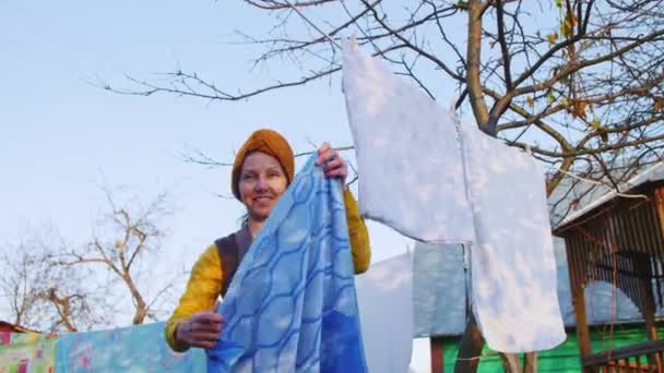 Glimlachende vrouw in gele hoofddoek hangt linnen in huis tuin — Stockvideo