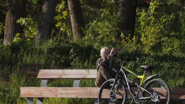 Молодая женщина на велосипеде снимает видео сидя на скамейке — стоковое видео