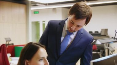 Takım elbiseli adam ofisteki bilgisayardaki kadınla konuşuyor.