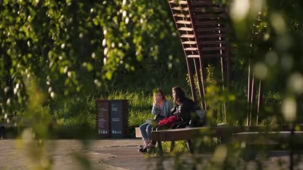 Unga kvinnor sitter på bänk nära trähus i grön park — Stockvideo