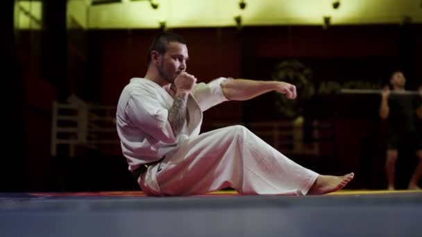 Hombre en kimono blanco hace abdominales crujidos patadas en el gimnasio — Vídeos de Stock