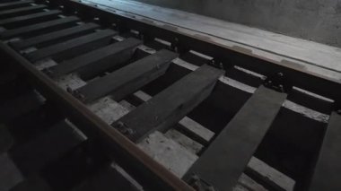 Tozlu ahşap yataklar loş beton çukurda ağır rayları destekliyor. Kasvetli endüstriyel atmosfer yeraltı inşaat sürecini vurguluyor