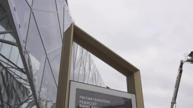 Cam üçgenlerle süslenmiş fütüristik tiyatro kareye hükmediyor. Posterli ilan panosu modern mimari ve sanatsal ruha zıt.