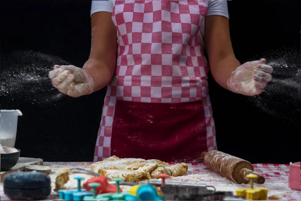 女の子は手をクラップし 暗い背景に生地の上に小麦粉を噴霧 エプロンのプロの女の子パン屋はクッキーを準備しています — ストック写真
