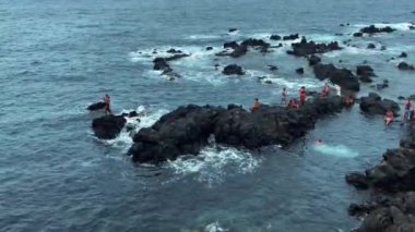 Üst Manzara. Bir grup insan, sahil kayalarının yanındaki çalkantılı bir denizde yüzüyor. Çarpışan dalgalarda aşırı yüzme