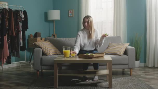 Ung flicka i vit tröja lägger glas apelsinjuice på bordet och biter pizza — Stockvideo