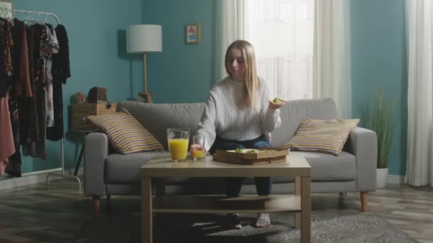 Молодая девушка в белом свитере держит пиццу и берет стакан апельсинового сока — стоковое видео