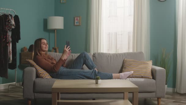 Chica joven acostada en el sofá y hablando de teléfono a través de enlace de vídeo — Vídeos de Stock