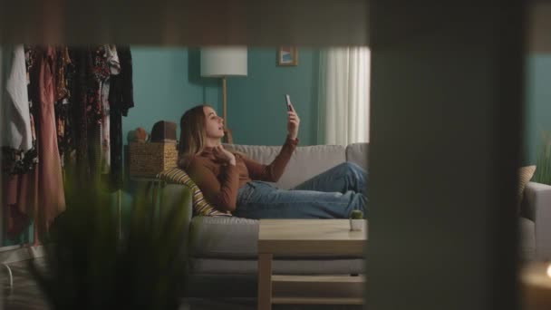 Молода дівчина лежить на дивані і розмовляє по телефону через відеозв'язок — стокове відео