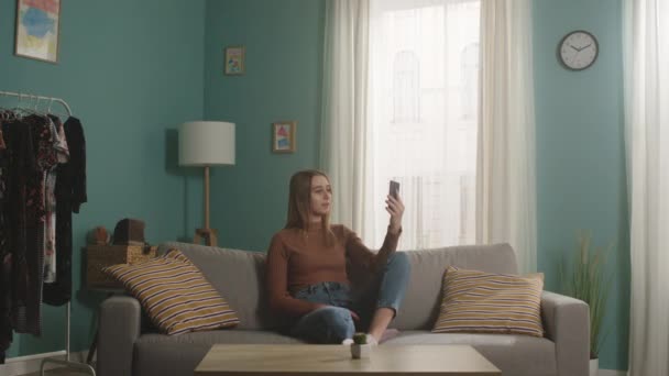 Young girl is sitting on a sofa and talking on the phone via video link — Stock Video