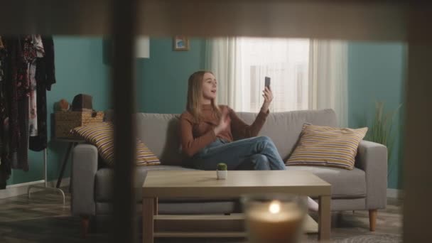 Jeune fille assise sur le canapé et téléphone parlant via lien vidéo — Video
