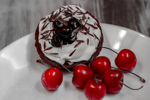Close up front view of black cake with white cream and cherry. Delicious the chocolate-rum cake decorated with milk cream and red cherry on a white plate on gray wooden background.